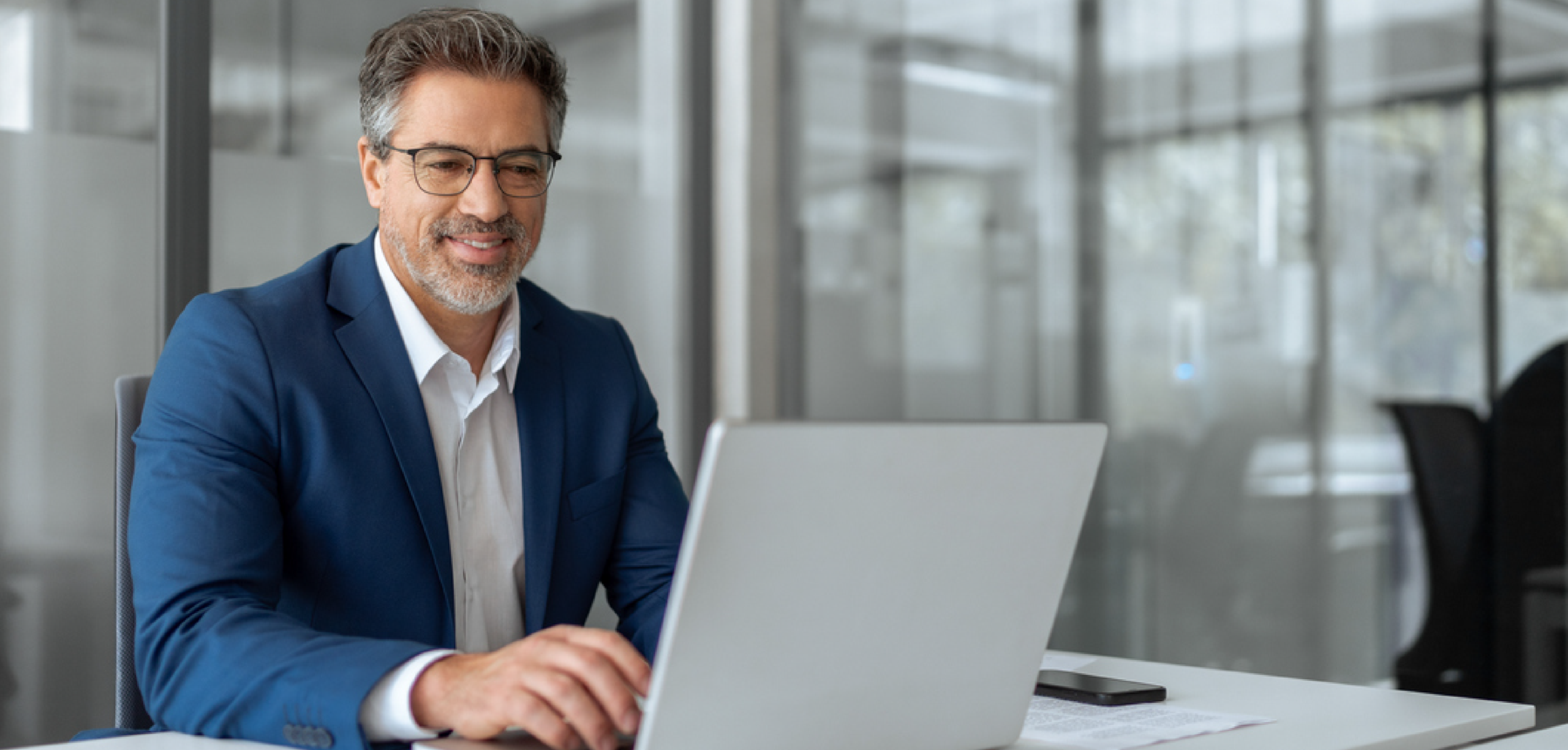 a man in glasses sitting at his desk and looking at his laptop